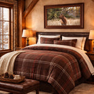 A rustic bedroom scene featuring a bed dressed in a brown and red plaid comforter set with matching pillows. Above the bed hangs a framed print of a bull elk in a field.