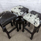 Three matching stools with dark wood legs and black-and-white cowhide upholstered seats, arranged on a carpeted floor against a white brick wall.