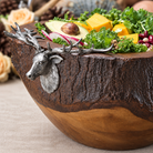 A rustic wooden salad bowl with a carved bull elk head and antler handle, filled with fresh greens, avocado, cheese cubes, red onion, and nuts, set on a neutral fabric surface.