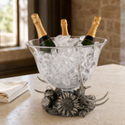 A clear glass champagne bucket filled with ice on a decorative silver base, with three champagne bottles placed inside, set on a tabletop in an indoor setting.