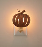A metal pumpkin-shaped night light with a warm glowing light inside, mounted on a white electrical outlet against a beige wall.