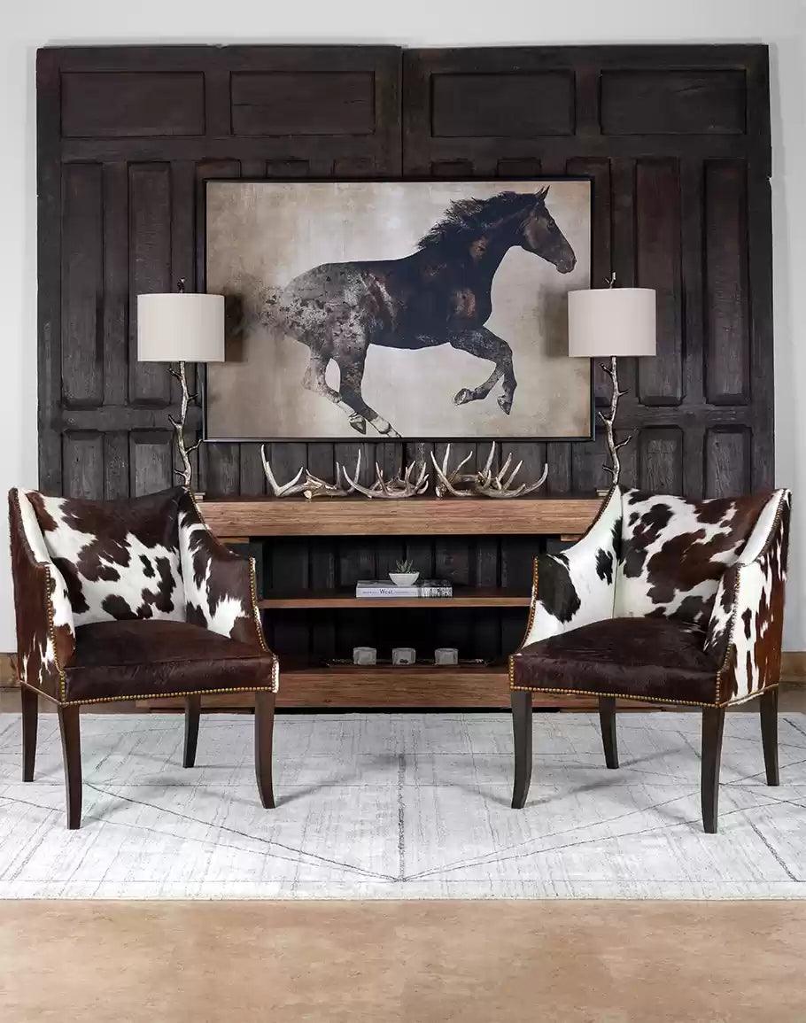 Cowhide chairs in sitting area with bronze lamps and antler decor, hickory console table and running horse art all on a light grey area rug - Your Western Decor