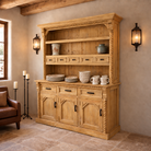 A large rustic wooden hutch or buffet cabinet with carved details, rope-textured trim, and multiple shelves and drawers, styled in a warm, rustic room with a brown leather chair, wall sconces, and Blue Mountain Brands Dinnerware