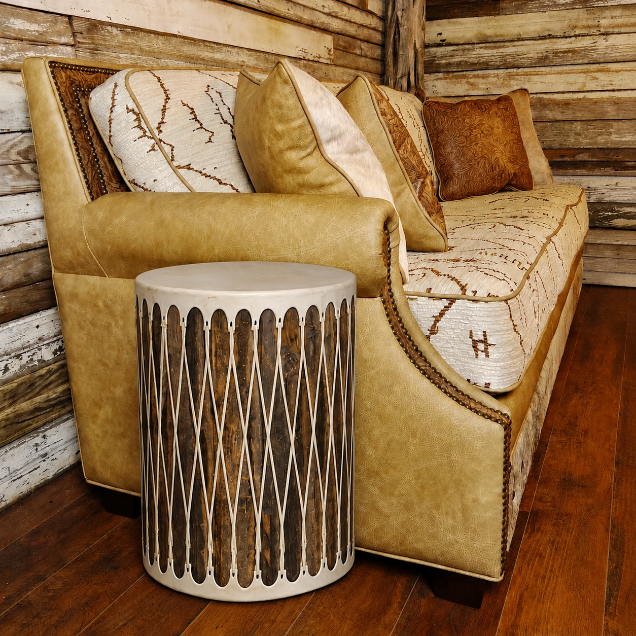 A rustic tan leather sofa with patterned cream and brown pillows, paired with a cylindrical side table featuring dark wood inlay and rawhide top and bottom with diamond lattice lacing, set against a wood-paneled wall and hardwood floor.