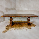 A rustic rectangular wooden dining table with turned block legs and a distressed finish, styled on a cowhide rug against a textured concrete wall.