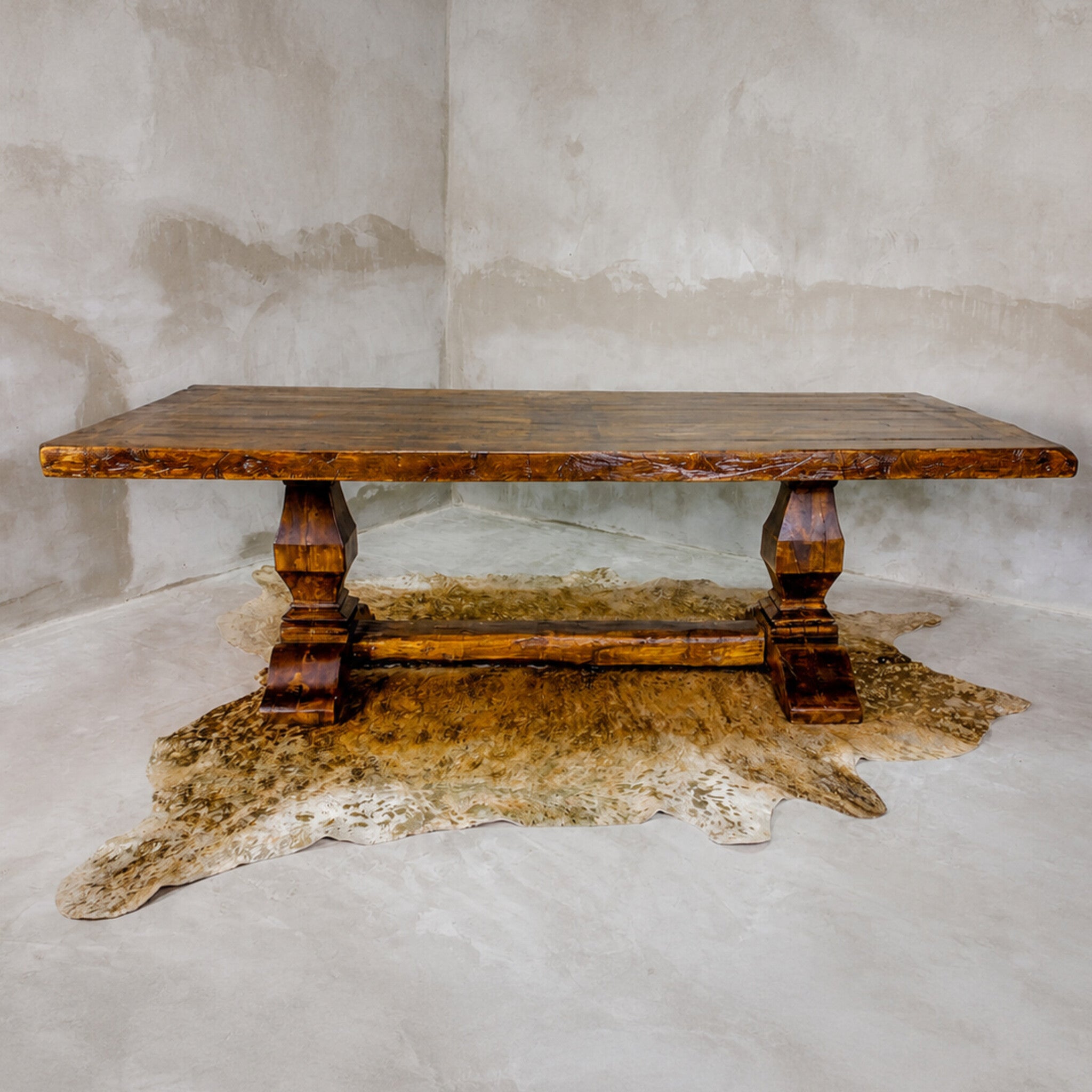 A rustic rectangular wooden dining table with turned block legs and a distressed finish, styled on a cowhide rug against a textured concrete wall.
