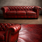 A classic red leather Chesterfield sofa with deep button tufting and brass nailhead trim, shown in a staged room setting with a matching leather rug in the foreground.
