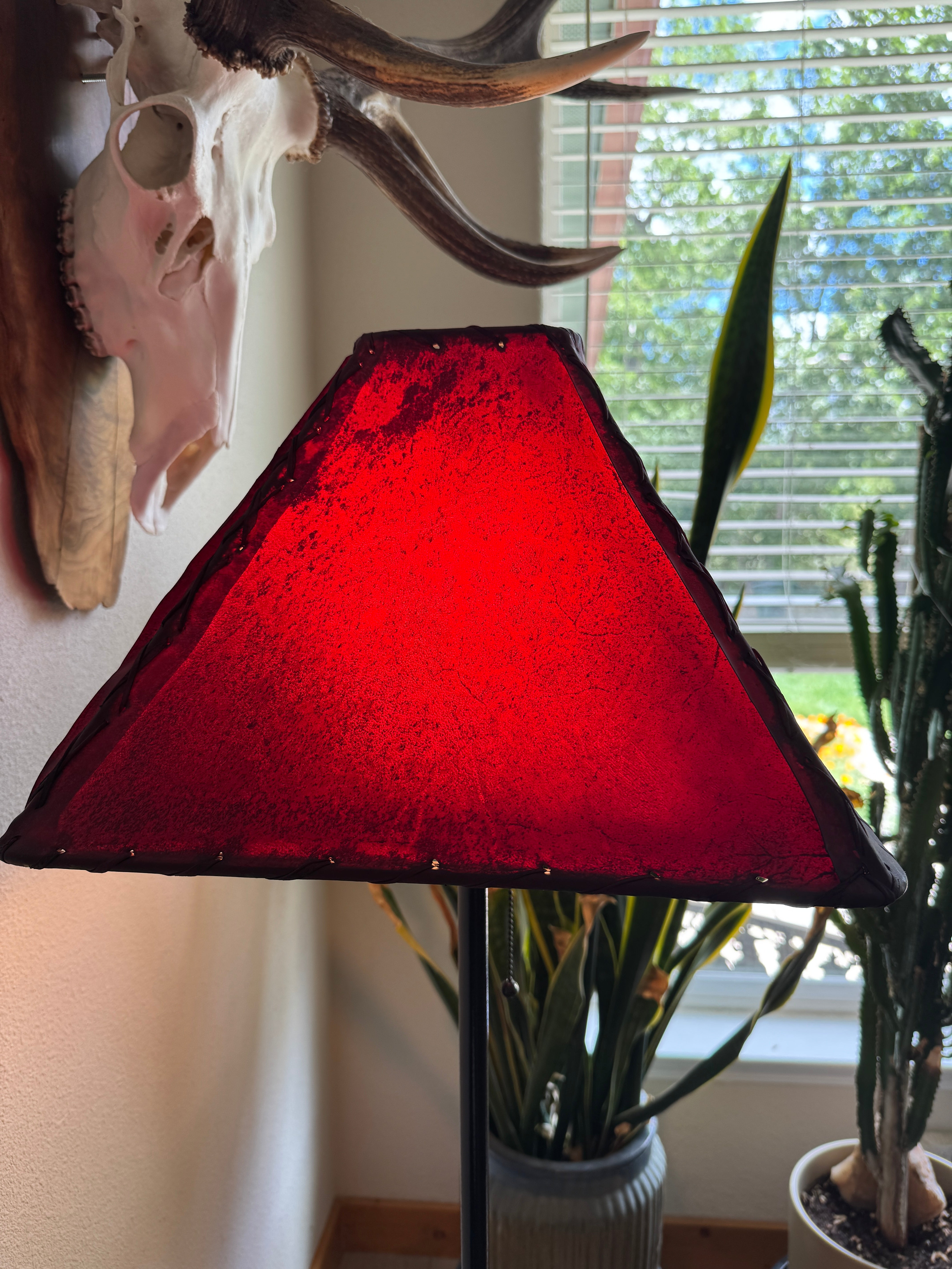 Red rawhide lampshade with light on, on a stand with a window and plants in the background