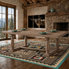 A rustic rectangular dining table with turned stone-like legs sits on a patterned southwestern-style area rug in a cabin-style dining room. The room has exposed wood beams, a stone fireplace, large windows with a mountain view, and a mounted longhorn artwork on the wall.