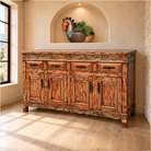 A rustic distressed wood sideboard cabinet with carved details, shown in a warm-toned room with a window, light wood floor, and an arched niche above holding a decorative horse-shaped planter.