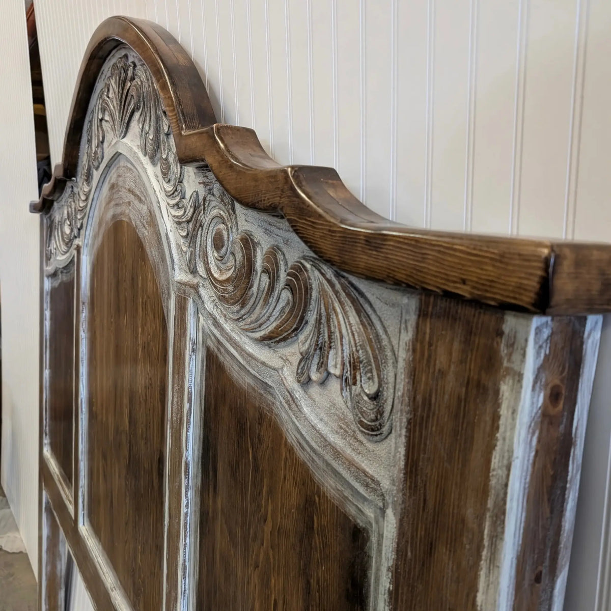Close-up of a rustic wooden headboard with ornate carved details and a distressed white paint finish showing through the wood grain.