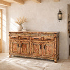 A rustic, distressed wooden sideboard cabinet with carved details, set in a warm, sunlit room with a textured plaster wall and a black wall sconce.