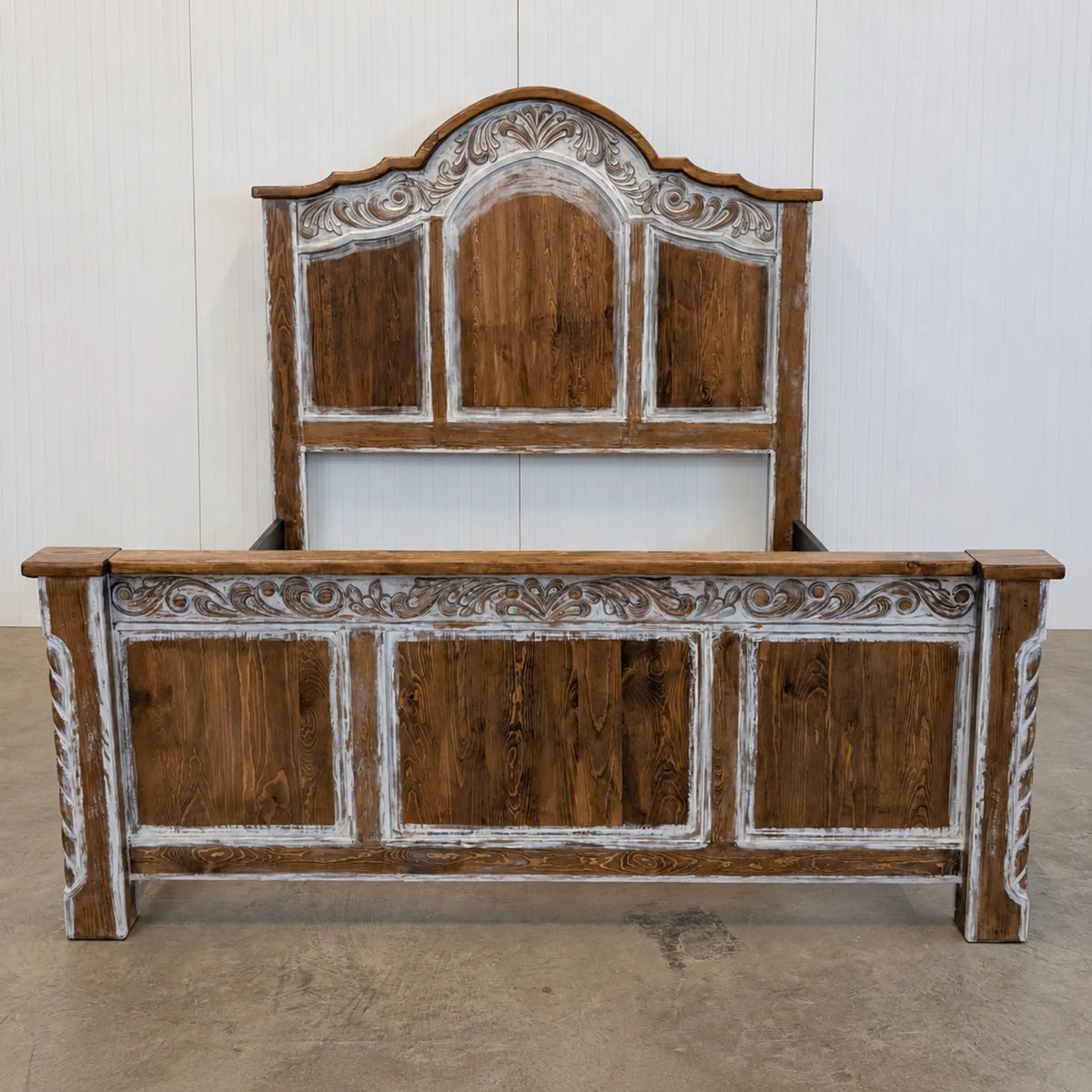 A rustic wooden bed frame with a tall matching headboard, featuring distressed brown wood panels and carved white accents, photographed in a warehouse setting.