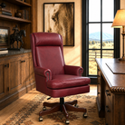 A high-back burgundy leather swivel office chair with nailhead trim and a dark wood five-star base, placed in a rustic home office setting with wood furniture, a window view, and wall art.
