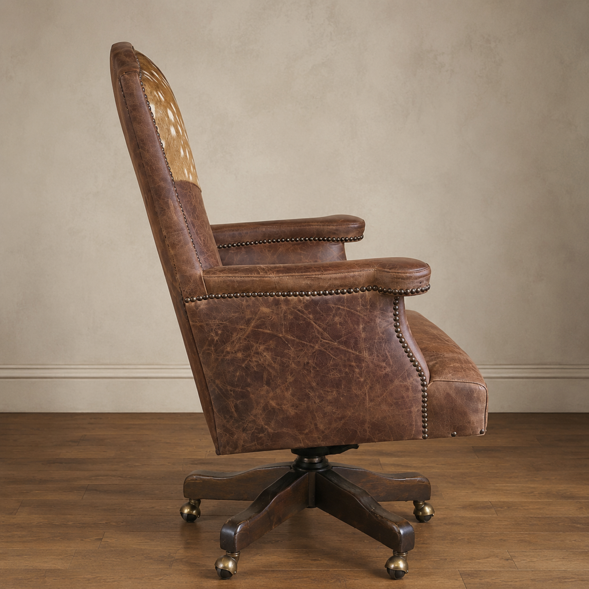 A brown distressed leather office swivel armchair with Axis hide yoke, nailhead trim and a dark wooden five-star base on a wood floor against a textured beige wall.