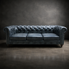 A dark blue tufted leather Chesterfield sofa with rolled arms and nailhead trim, photographed against a dark textured studio backdrop.