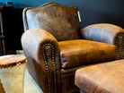 A brown leather armchair with textured crocodile-patterned backrest panels and brass nailhead trim, shown in a furniture setting with a matching ottoman partially visible.