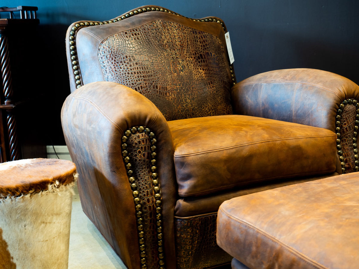 A brown leather armchair with textured crocodile-patterned backrest panels and brass nailhead trim, shown in a furniture setting with a matching ottoman partially visible.