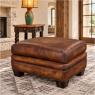 A square brown leather ottoman with a distressed finish and dark wooden legs, shown in a styled living room setting on a patterned rug next to a side table with a lamp.