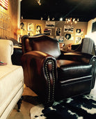 A dark brown leather club chair with nailhead trim, photographed in a furniture showroom with other chairs, wall art, and lighting fixtures visible in the background.