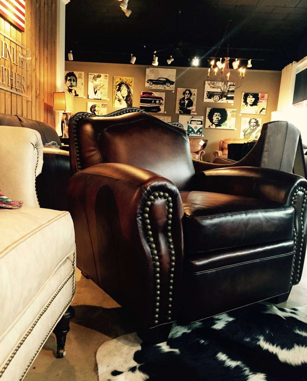 A dark brown leather club chair with nailhead trim, photographed in a furniture showroom with other chairs, wall art, and lighting fixtures visible in the background.