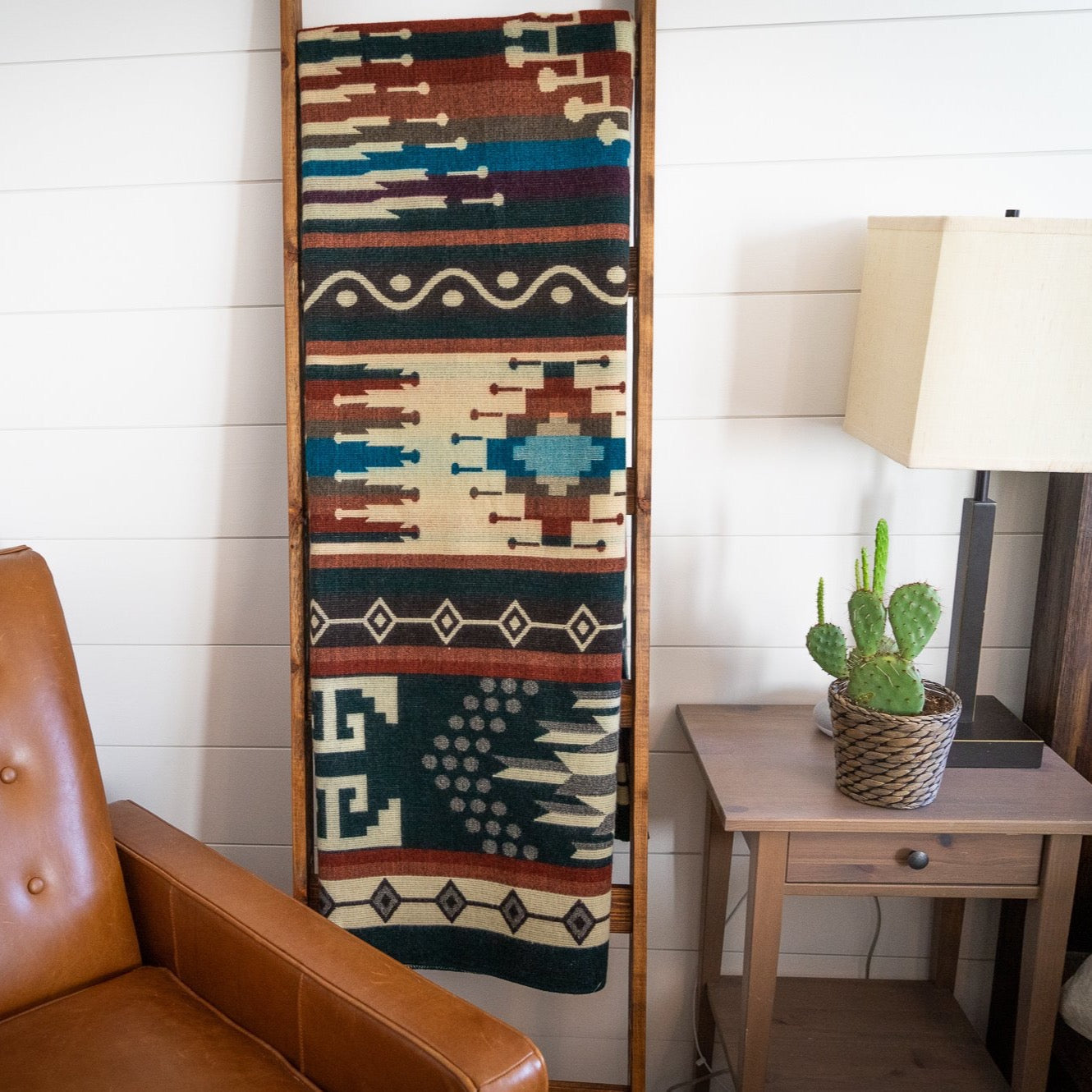Decorative Alpaca blanket with geometric pattern on a wooden ladder, brown leather chair, and small table with lamp and cactus.