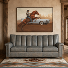 A framed western-style wall art print hangs above a gray leather sofa in a rustic room with wood beams and a patterned rug. The artwork depicts a cowboy riding a brown horse in front of a light blue classic car.