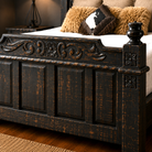 A dark carved wooden bed frame with distressed brown and black finish, shown in a bedroom setting with white bedding and decorative pillows.
