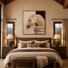 A rustic mountain-view bedroom with a large framed horse portrait centered above the bed. The room has warm beige walls, two matching nightstands with lit lamps, curtains, a bench at the foot of the bed, and mountain scenery visible through the windows.