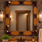 A rustic rectangular bathroom mirror with a decorative frame made of dark brown wood, cowhide patches, and silver studs, mounted on a stone wall with wall sconces and a sink below.