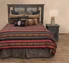 A rustic western-style bedding set on a bed, featuring a striped pattern in red, brown, black, and beige with a Southwestern geometric accent pillow. The bed is styled with a dark wood headboard and a matching dark wood nightstand with a lamp, set against a textured beige wall and wood floor.