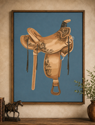 A framed art print featuring a detailed illustration of a tan leather western saddle with floral engravings, set against a muted blue background. The print is displayed on a textured beige wall above a wooden shelf, flanked by a small bronze horse figurine on the left and a potted plant on the right.