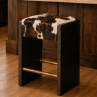 A rustic wooden stool with a black frame, a cowhide upholstered seat, and brass-colored metal footrests, set in a warm wood-paneled room.