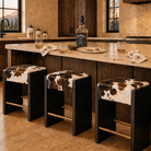 Three modern western bar stools with black wood frames and cowhide seats sit at a kitchen island. A Jack Daniel's bottle and glasses are on the counter, with a rustic kitchen interior in the background.
