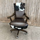 A rustic office chair with a dark brown faux-leather seat and back, featuring cowhide-patterned accents on the headrest and lower seat, mounted on a dark wood five-star swivel base with casters.