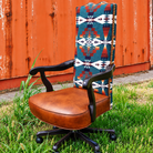 A high-back office chair with a brown leather seat and black swivel base, featuring a tall backrest upholstered in a Southwestern-style geometric patterned fabric, set outdoors on grass in front of a rusted orange metal fence.