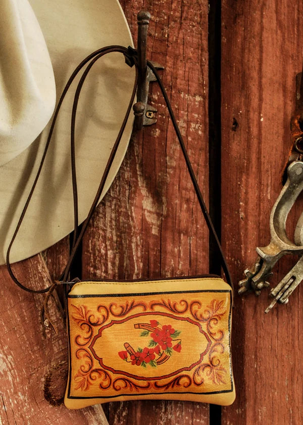 A small tan leather crossbody leather bag with hand-painted red floral and scroll designs, hanging from a rustic metal hook on a weathered red wooden wall. A cream-colored cowboy hat is draped over the left side, and a metal spurs ornament is visible on the right.