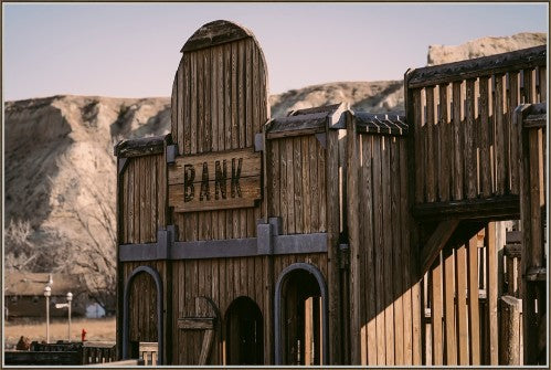 Western ghost town bank canvas art by Adam Mowrey featuring a weathered wooden facade, bank sign, and rugged hills in an oak-look floater frame. Made in the USA
