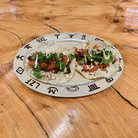 Two soft corn tacos topped with diced tomatoes, cilantro, and a white sauce, served on a beige plate with dark brown ranch brands and rope images, placed on a wooden table.