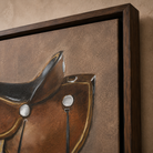 A close-up of framed wall art showing a painted brown leather saddle against a textured brown background, set in a dark wood frame against a beige wall.
