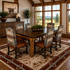 A rustic dining room set featuring a dark wood rectangular table and six matching high-back chairs with patterned upholstery and brown leather seats, arranged on a southwestern-style area rug in a cabin-style room with large windows.