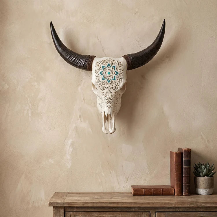 A decorative mounted animal skull with long curved dark horns and an ornate cream skull face featuring a blue and teal geometric mandala pattern, displayed on a textured beige wall above a wooden console table with books and a small succulent.