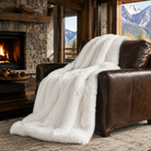 A fluffy white faux fur throw blanket is draped over a brown leather armchair in a rustic cabin living room with a stone fireplace and mountain windows.