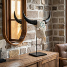 A decorative animal skull with long curved black horns and a white face, mounted on a black stand on a wooden console table in a rustic room with a stone wall and a large oval mirror.