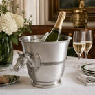 A luxury pewter champagne bucket with a horse head handle and rope detail, filled with ice and a bottle of champagne on a dining table, with two filled champagne flutes, plates, and a vase of white flowers nearby.