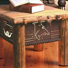 A rustic wooden table with a thick log-top surface and log-style legs, featuring cutout western motifs on the front panel. Books rest on top, and the table is shown in a warm indoor setting with wood flooring.