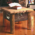 A rustic log-style end table with metal cutout bronc and western motifs, shown in a cozy room setting with a fireplace, books on top, and a wooden floor.