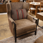A brown wingback armchair with dark wood legs and nailhead trim, shown in a furniture showroom. The chair is fitted with a matching brown striped accent pillow featuring red, blue, and beige stripes.