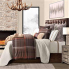 Brown plaid comforter set layered with light grey quilt, knitted euro shams, and faux fur throws with fireplace and antler chandelier